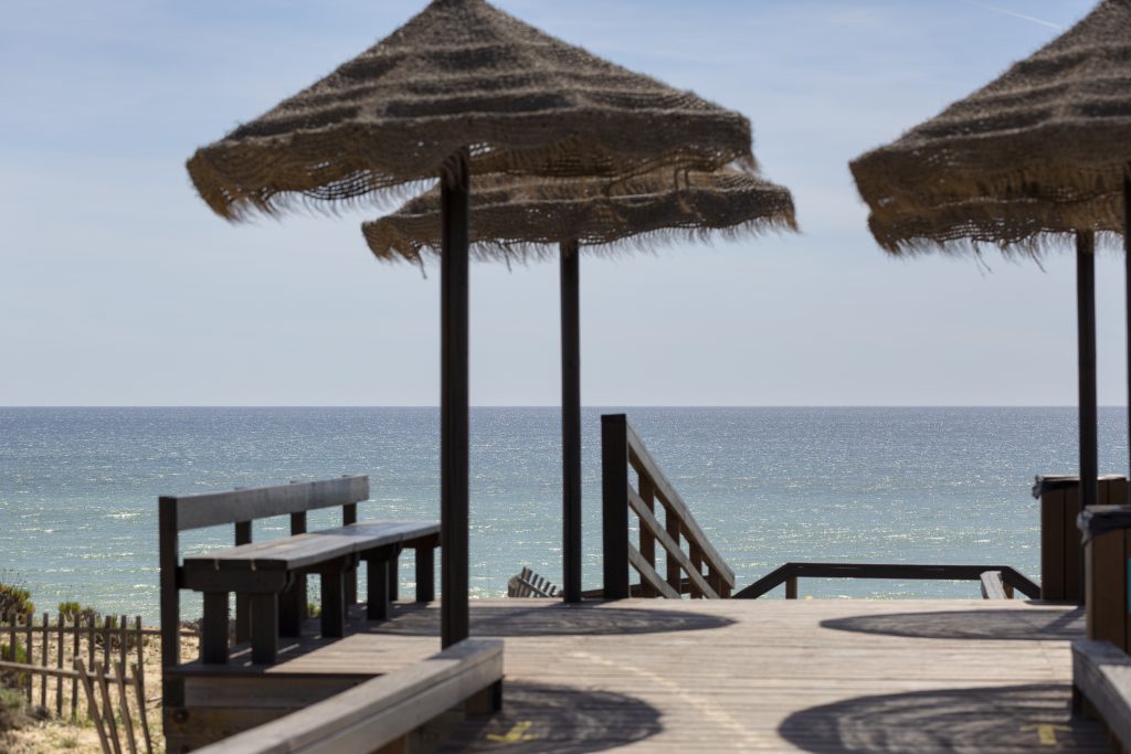 Wooden pathway leading towards the calm blue Atlantic Ocean, featuring thatched beach umbrellas and a wooden bench for beach access and relaxation.