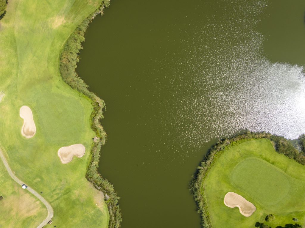 Top-down aerial view of a meticulously maintained green golf course, showcasing sand bunkers and a large, dark water hazard or lake.