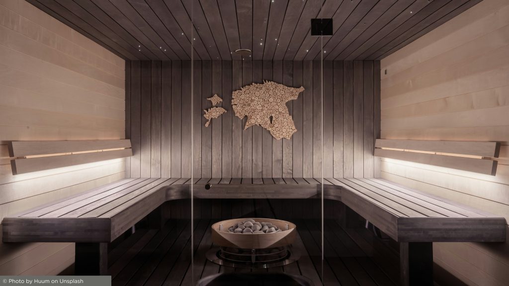 Modern sauna with wood benches, a log-map of Estonia on the wall, central stone stove, soft lighting, and sleek glass panels for a serene ambiance.
