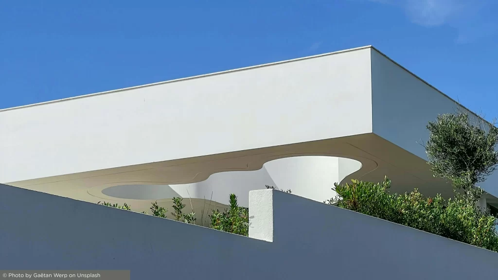 Composição abstrata apresentando uma parede branca com uma perspetiva parcial de um jardim paisagístico e cobertura da habitação, refletindo sofisticação e pureza de linhas arquitetónicas.